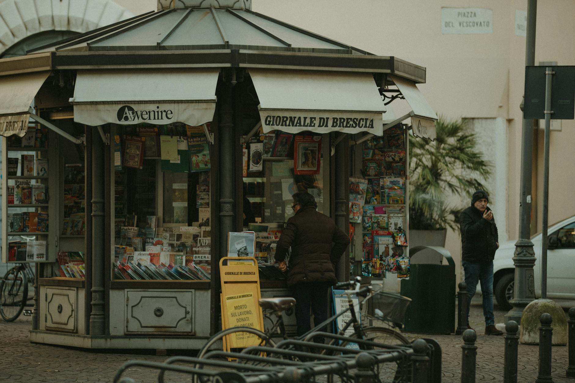 Street view of a newspaper kiosk in Brescia, Italy, featuring daily publications and passerby.