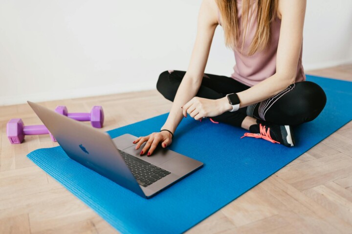 Faceless female in activewear sitting on blue yoga mat on floor and browsing laptop